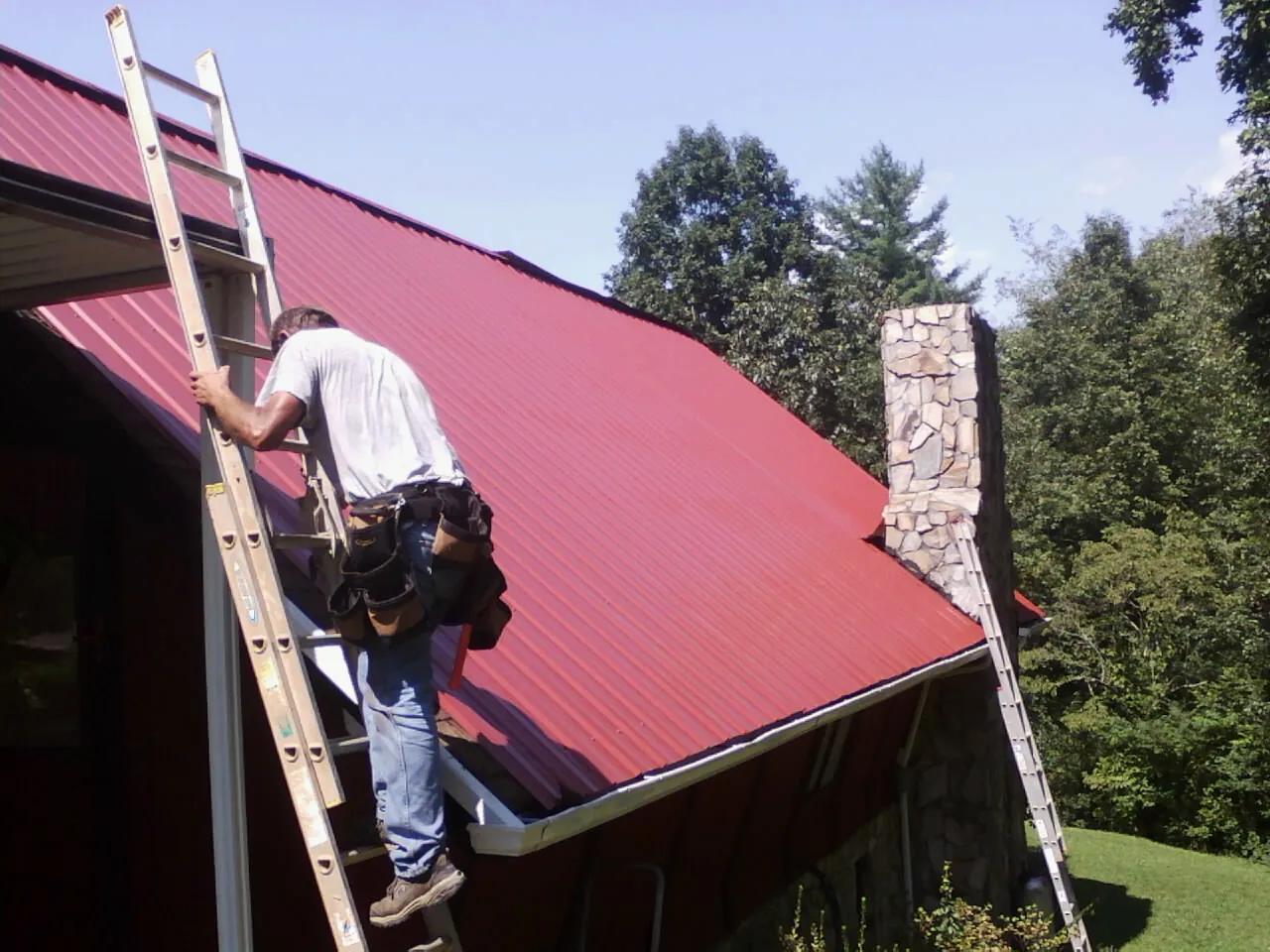 Roofer on a ladder beside a red metal roof and stone chimney — Soffit & Fascia Repair by Roofing Brothers in Boothbay
